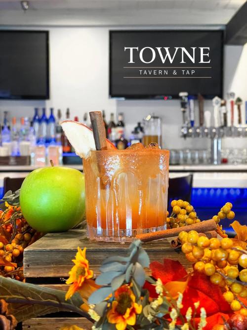 A cocktail garnished with a cinnamon stick and apple slice, surrounded by autumn decor and a green apple.