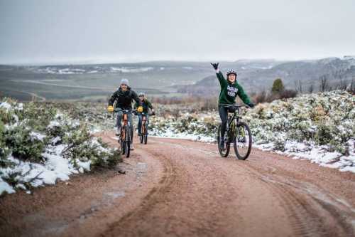 A group of four mountain bikers riding on a snowy dirt trail, with one person joyfully waving.