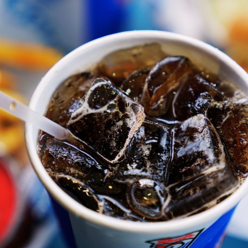 A close-up of a cup filled with dark soda and ice cubes, with a straw partially visible.