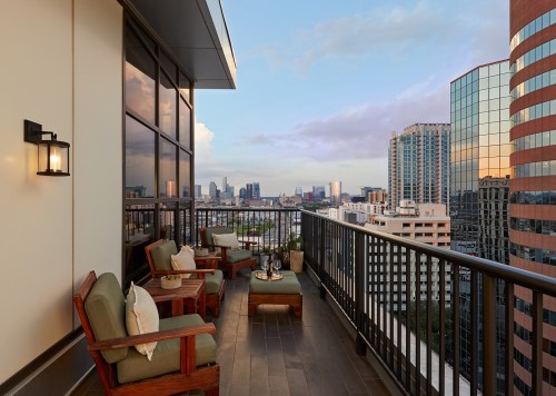 A balcony with seating and a view of a city skyline at sunset, featuring modern buildings and a clear sky.