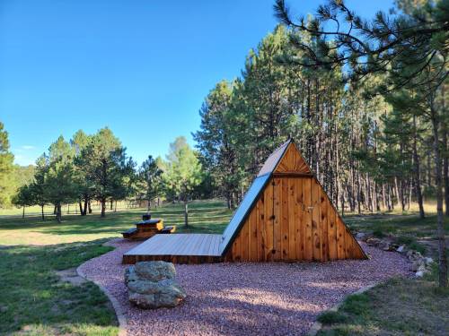 A wooden A-frame structure sits on a gravel area surrounded by trees in a sunny outdoor setting.