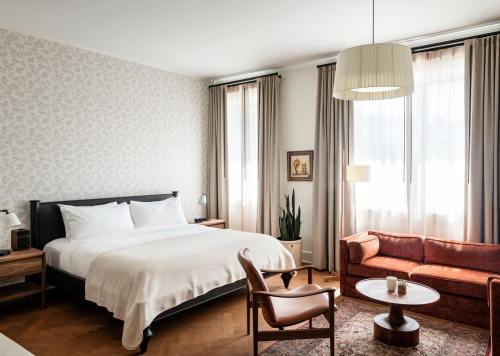 Cozy hotel room with a large bed, armchair, coffee table, and natural light from tall windows. Elegant decor and plants.