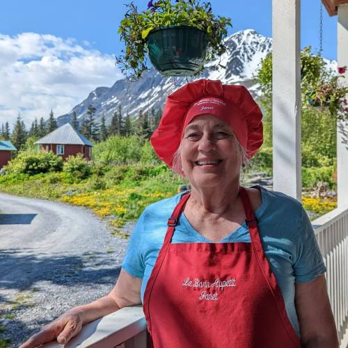 Smiling woman in a red chef hat and apron stands on a porch with mountains and flowers in the background.