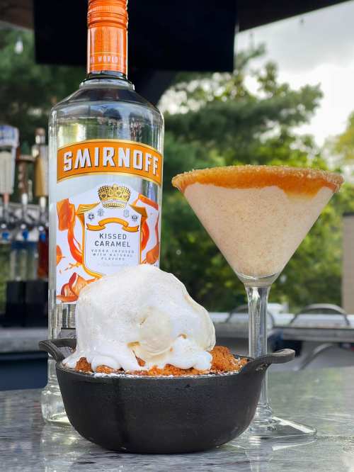 A bottle of Smirnoff Kissed Caramel vodka next to a caramel martini and a dessert with ice cream in a cast-iron dish.