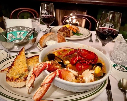 A bowl of seafood stew with crab and lobster, accompanied by garlic bread, wine, and side dishes on a dining table.
