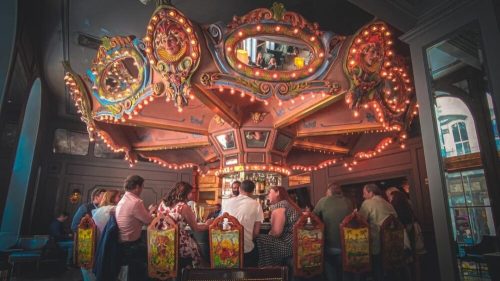 A vintage carousel-themed bar with patrons enjoying drinks under warm lights and ornate decorations.