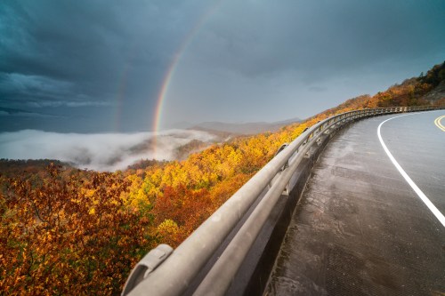 A winding road curves along a colorful autumn landscape with a double rainbow over misty mountains.