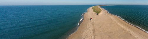 Aerial view of a sandy peninsula extending into a calm blue sea, with a vehicle parked on the shore.