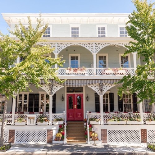 A charming light blue building with white trim, featuring a red door and lush greenery around the entrance.