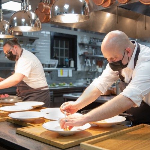 Two chefs in a kitchen, wearing masks, carefully plating dishes on wooden boards. Copper pots hang in the background.