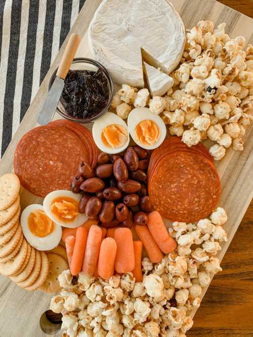 A wooden platter with cheese, salami, olives, boiled eggs, carrots, crackers, and popcorn arranged artfully.