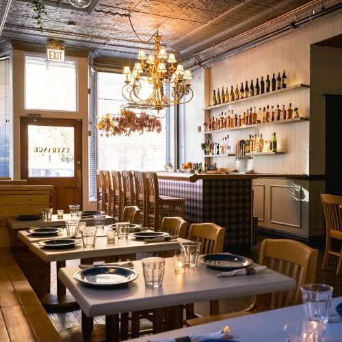 Cozy restaurant interior with wooden tables, a chandelier, and a bar lined with bottles, illuminated by natural light.