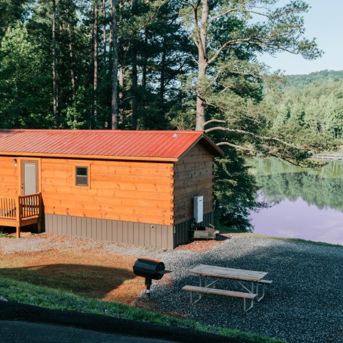 A wooden cabin with a red roof by a calm lake, surrounded by trees and a picnic area with a grill.