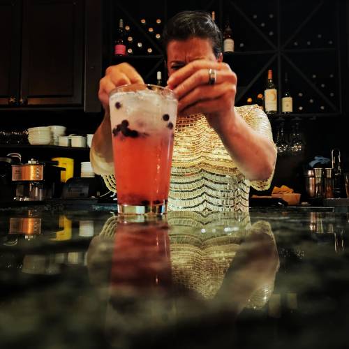 A person in a shiny top prepares a colorful drink at a bar, with reflections on the countertop.