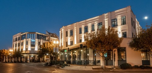 A well-lit urban scene at dusk featuring modern buildings and trees lining the street.