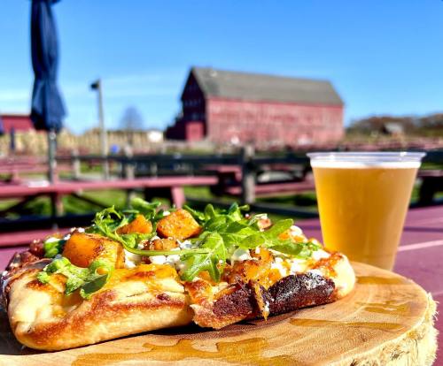 A flatbread topped with cheese, arugula, and squash, alongside a pint of beer, with a red barn in the background.