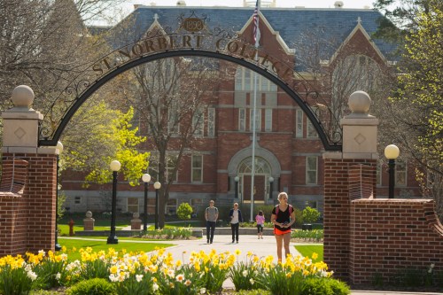 A sunny campus scene at St. Norbert College, featuring blooming flowers and people walking under an archway.