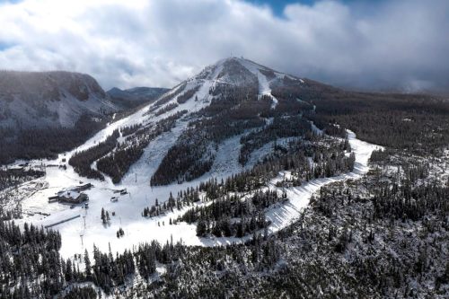 Aerial view of a snow-covered mountain with ski trails and a lodge surrounded by evergreen trees under a cloudy sky.