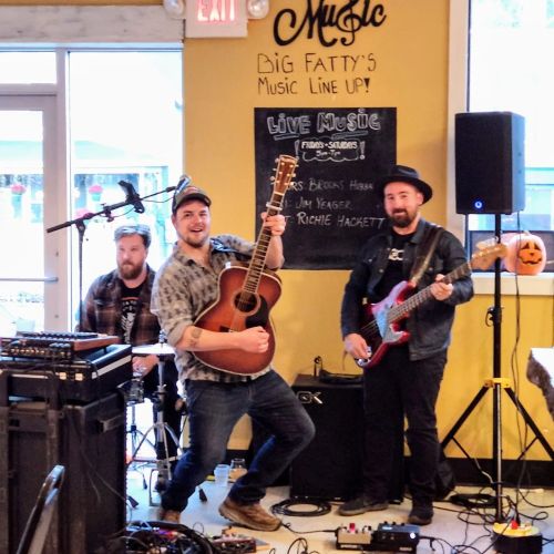 A lively band performs in a cozy venue, with a guitarist, bassist, and drummer on stage, surrounded by musical equipment.