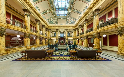 Luxurious hotel lobby with ornate architecture, marble columns, and a grand staircase under a glass ceiling.