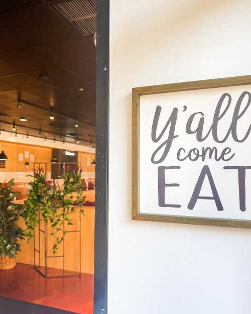 A sign reading "Y'all come EAT" next to a doorway leading into a cozy restaurant with plants.