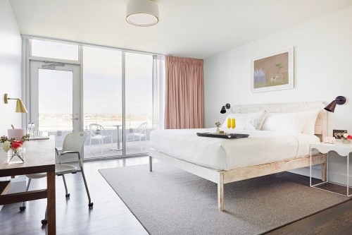 Bright hotel room with a large window, modern decor, a bed, desk, and balcony access. Soft colors and natural light.
