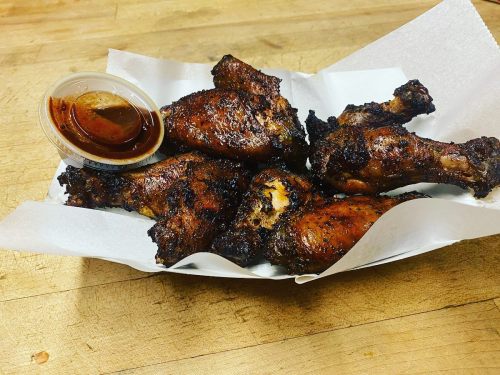 A basket of crispy, grilled chicken wings with a small cup of barbecue sauce on a wooden surface.