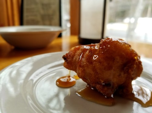 A close-up of a fried dessert drizzled with syrup on a white plate, with a blurred background of a table setting.