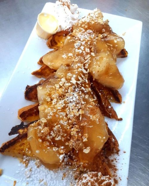 A plate of dessert featuring sliced, caramelized apples on toasted bread, topped with whipped cream and powdered sugar.