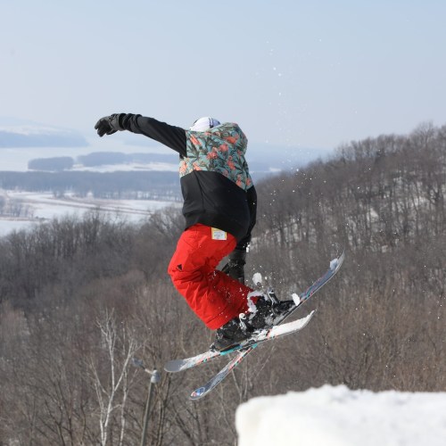 A skier in a colorful jacket and red pants jumps off a snowy slope, with trees and hills in the background.