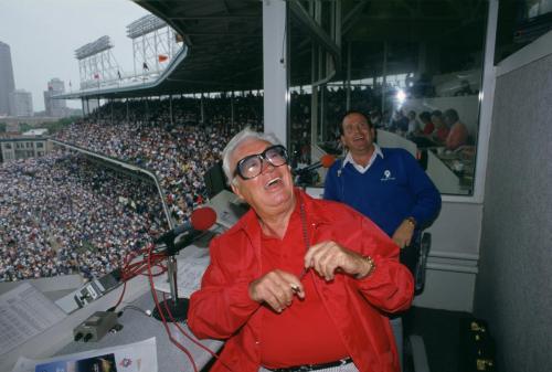 Two men in a broadcast booth at a baseball stadium, with a large crowd visible in the background. One is laughing.