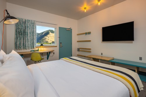 A cozy hotel room featuring a bed, desk, TV, and a window with a mountain view. Bright and modern decor.
