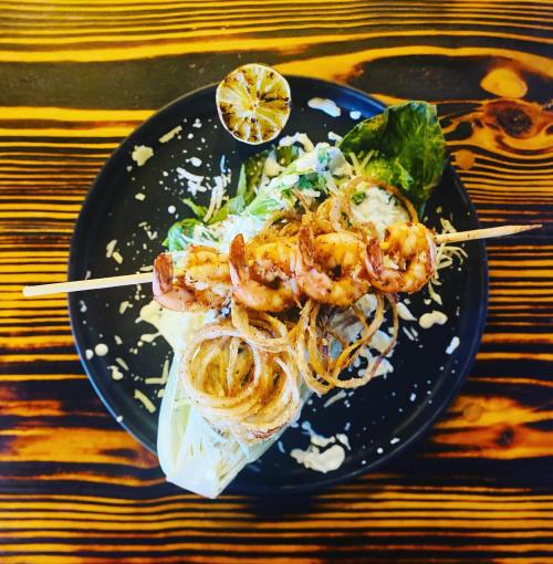 Grilled shrimp skewers on a bed of lettuce, garnished with crispy onions and a lime wedge on a black plate.
