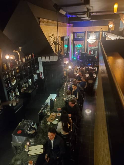 A dimly lit bar scene with patrons seated at the counter and tables, surrounded by shelves of bottles and elegant decor.