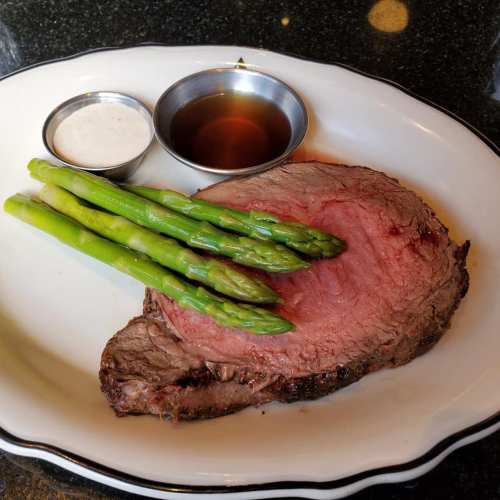 A plate with a slice of prime rib, asparagus spears, and two dipping sauces on the side.