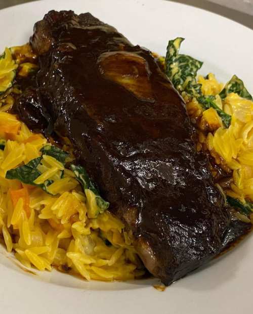 A plate of tender meat glazed in a dark sauce, served over a bed of colorful orzo and greens.