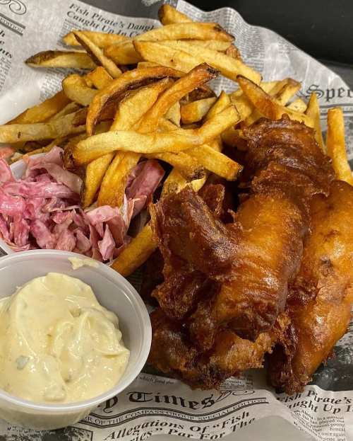 A plate of crispy fried fish, golden fries, pink coleslaw, and a small cup of creamy sauce, served on newspaper.