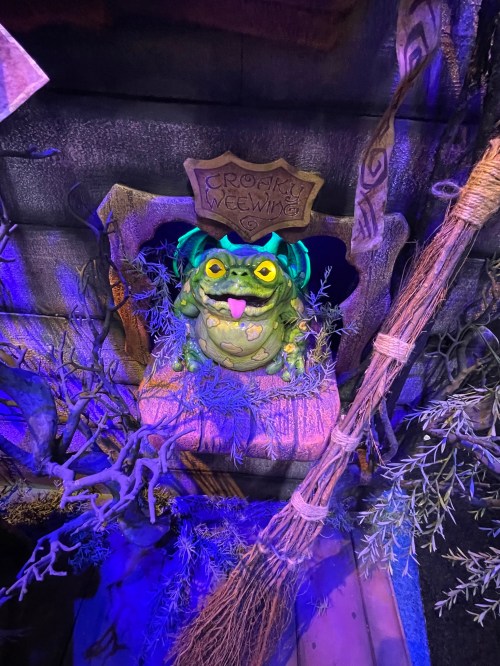 A whimsical green frog statue with yellow eyes sits in a stone alcove, surrounded by dark foliage and a broom.