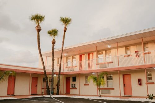 A pastel-colored motel with palm trees, featuring a two-story structure and red doors under a cloudy sky.
