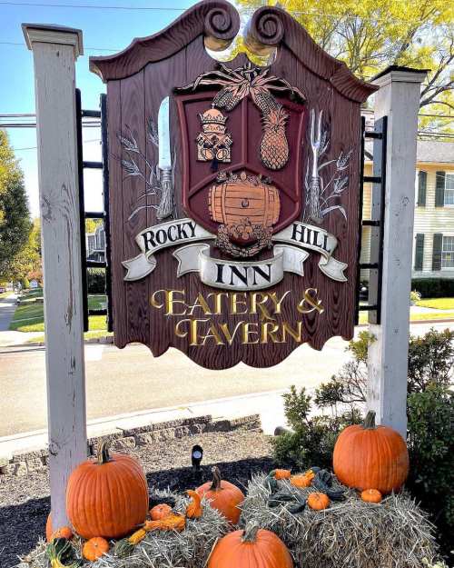 Sign for Rocky Hill Inn Eatery & Tavern, featuring a coat of arms, pumpkins, and hay bales in a fall setting.