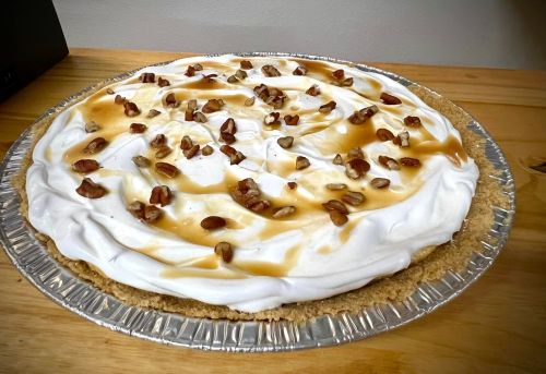 A creamy pie topped with whipped cream, caramel drizzle, and chopped nuts in a silver pie crust on a wooden surface.