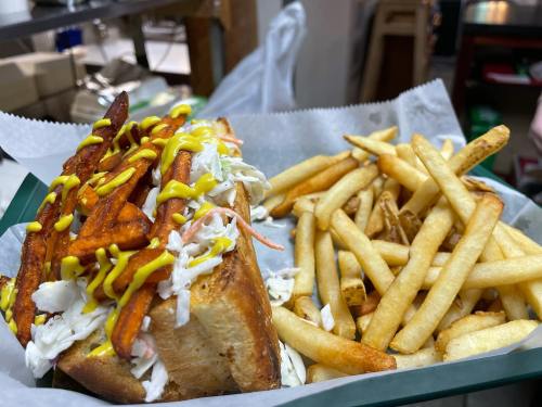 A sandwich topped with coleslaw and sweet potato fries, served with a side of golden French fries and mustard drizzle.
