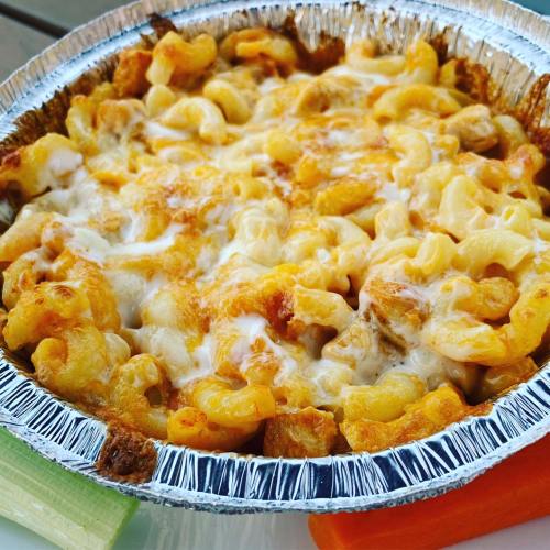 A baked macaroni and cheese dish in a foil container, topped with melted cheese, served with celery and carrot sticks.
