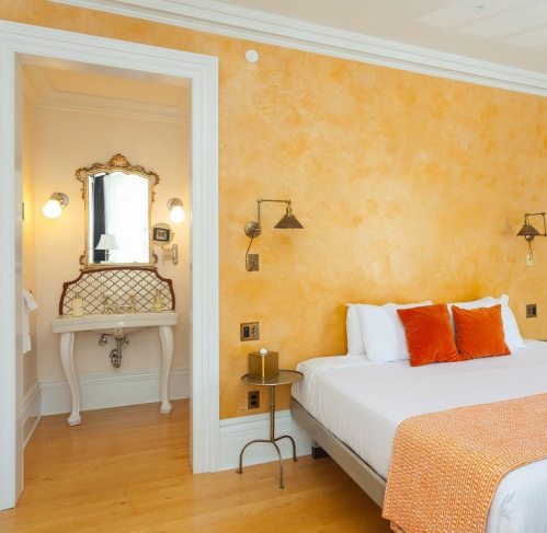 Cozy hotel room with orange walls, a bed with orange pillows, and an adjoining bathroom with a vintage mirror.