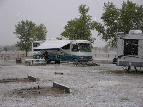 An RV parked in a snowy campground, with snow falling and trees in the background. Picnic tables are visible nearby.