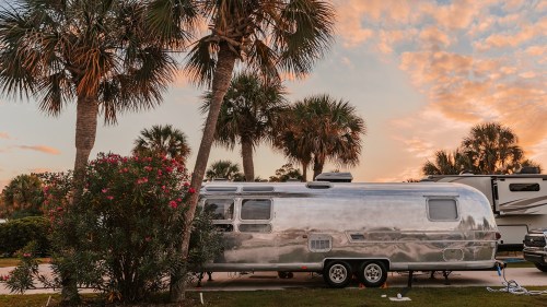 A shiny silver Airstream trailer parked among palm trees at sunset, with colorful flowers in the foreground.