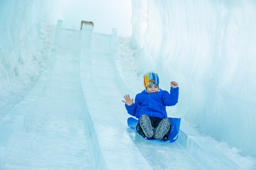 A child in a blue jacket sleds down a snowy slide, surrounded by icy walls.