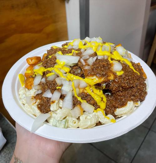A hand holding a plate of macaroni salad topped with chili, onions, and mustard.