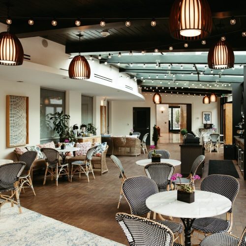 A stylish indoor café with wooden floors, modern lighting, and tables set for dining, featuring plants and cozy seating.