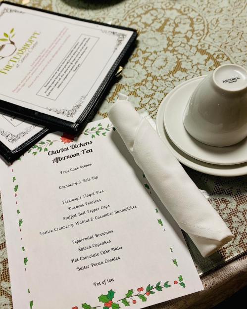 A table setting for afternoon tea featuring a menu, a teacup, and a neatly rolled napkin on a lace tablecloth.